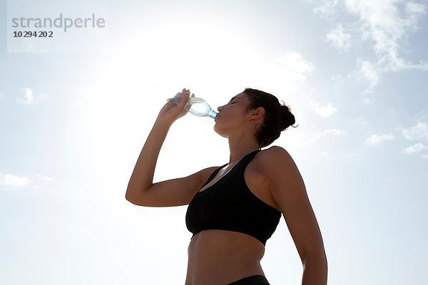 Niedriger Blickwinkel auf den blauen Himmel und die erwachsene Frau  die Wasser in Flaschen trinkt.