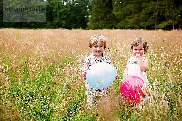 Bruder und Schwester im hohen Gras halten Ballon und schauen lächelnd in die Kamera.