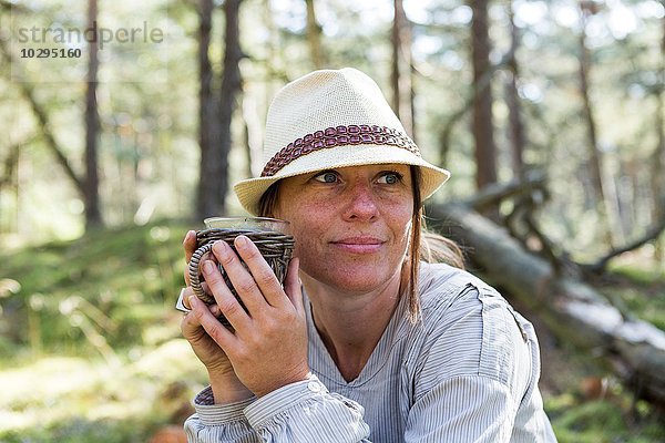 Reife Frau trägt Filzhut und trinkt Tee im Wald.