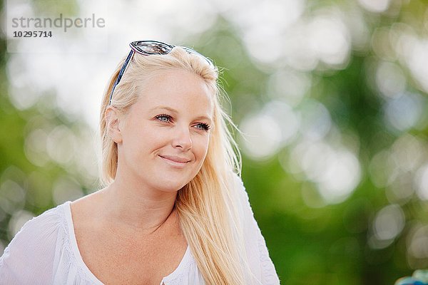 Porträt einer langhaarigen blonden jungen Frau mit Sonnenbrille auf dem Kopf  die lächelnd wegsieht.