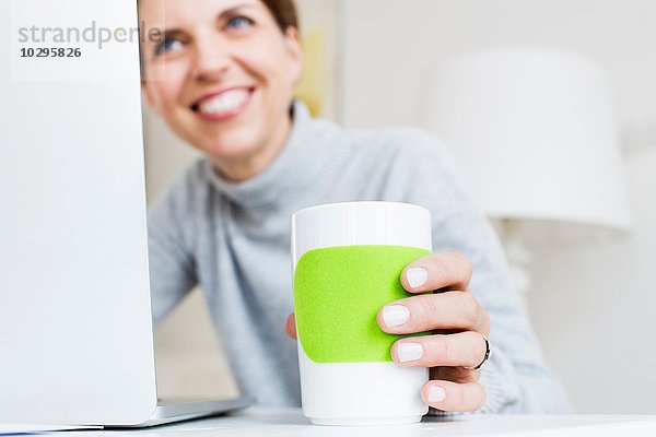 Reife Frau am Laptop hält Tasse lächelnd  Fokus auf Vordergrund