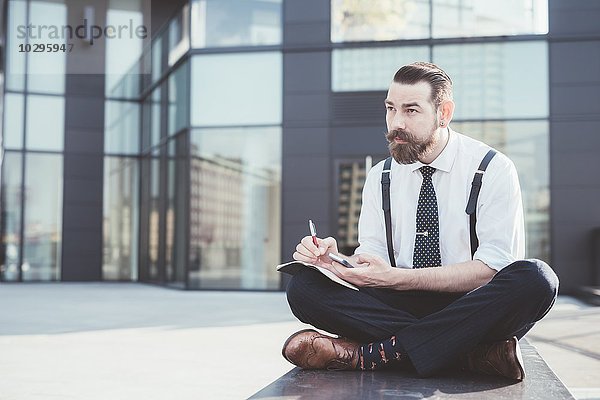 Stilvoller Geschäftsmann sitzt im Kreuzbein und macht Tagebuchnotizen vom Smartphone außerhalb des Büros.