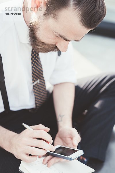 Geschäftsmann sitzt im Kreuzbein und macht Tagebuchnotizen vom Smartphone außerhalb des Büros.