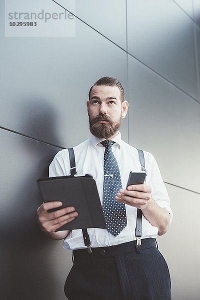 Stilvoller Geschäftsmann mit Smartphone und digitalem Tablett  das sich an die Bürowand lehnt