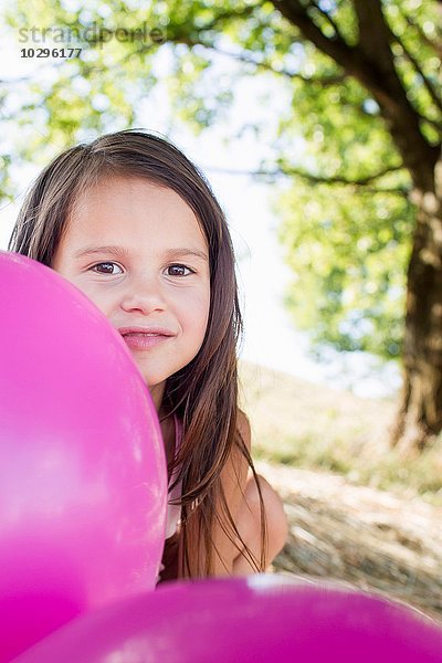 Porträt eines Mädchens  das sich hinter rosa Luftballons im Park versteckt.