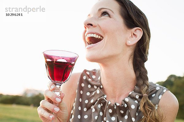 Reife Frau lacht  während sie im Park ein Glas Rotwein trinkt.