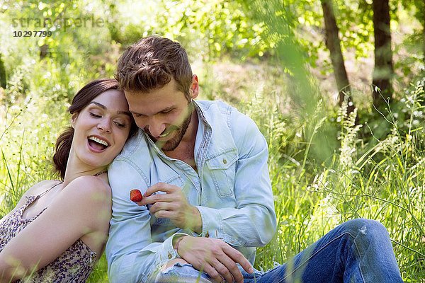 Junges Paar sitzt Rücken an Rücken auf Gras und hält Erdbeere lächelnd.