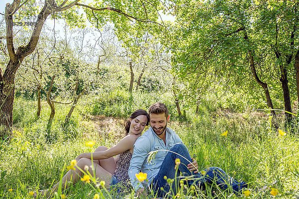 Junges Paar sitzt Rücken an Rücken auf Gras und schaut lächelnd nach unten.
