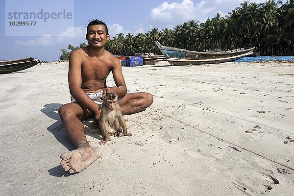 Einheimischer Mann sitzt mit einem Affen an der Leine am Strand des Fischerdorfes Ngapali  hinten Fischerboote  Thandwe  Rakhine-Staat  Myanmar  Asien