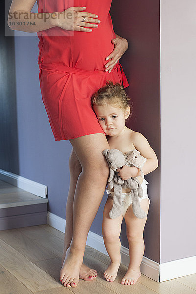 Kleines Mädchen klammert sich an das Bein ihrer schwangeren Mutter mit beunruhigendem Gesichtsausdruck.