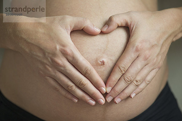 Schwangere Frau macht Herzform mit Händen um den Bauchnabel herum