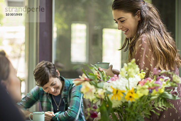 Drei sprechende und lachende Frauen  die um einen Tisch saßen. Großer Blumenstrauß in einer Vase.