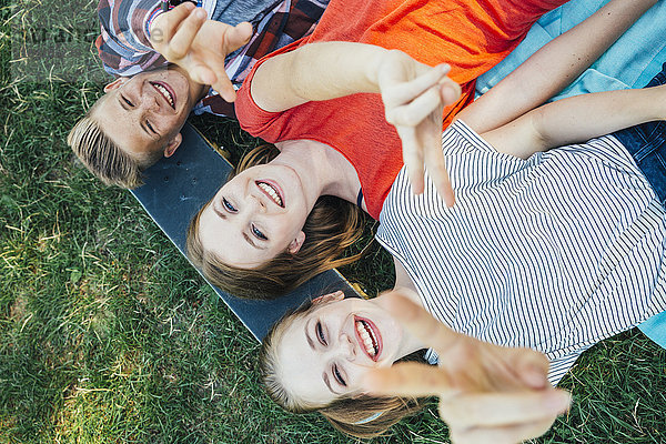 Drei glückliche Teenager-Freunde liegen auf dem Skateboard auf der Wiese und machen ein Siegeszeichen.