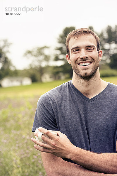 Ein junger Mann mit einer Kaffeetasse in der Hand steht auf einem Feld an einem See.