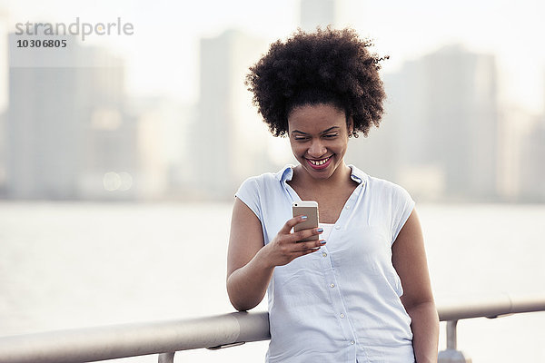 Eine Frau lehnt an einem Geländer am Wasser und überprüft ihr Handy