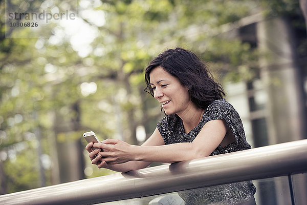 Eine Frau lehnt an einem Geländer in einem Stadtpark und überprüft ihr Handy