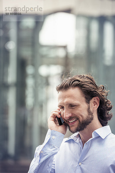 Ein Mann mit Bart  der auf einer Straße in der Stadt mit einem Handy telefoniert.
