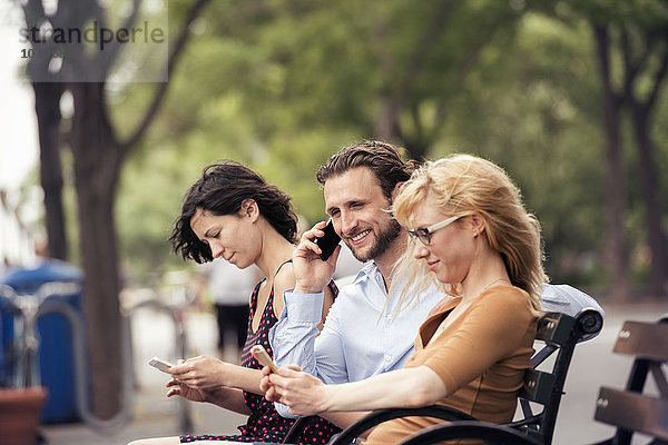 Ein Mann und zwei Frauen sitzen auf einer Parkbank und überprüfen ihre Telefone  eine telefoniert.
