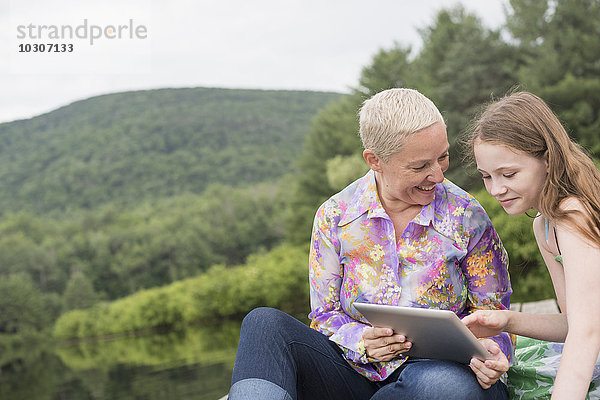 Eine Frau und ein junges Mädchen zusammen auf einem Dock am See mit einem digitalen Tablet.