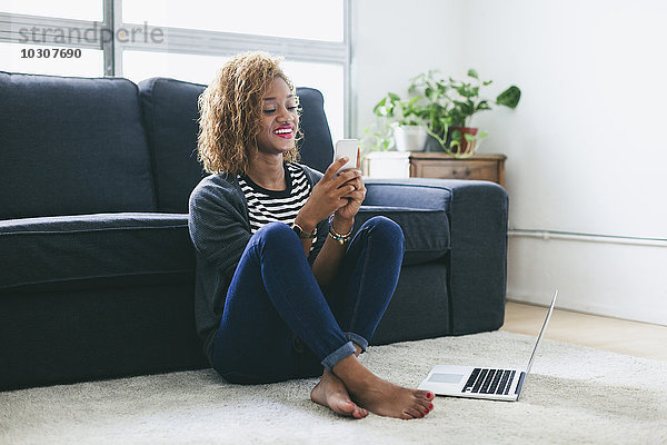 Lächelnde junge Frau sitzt auf dem Teppich des Wohnzimmers und schaut auf ihr Smartphone.