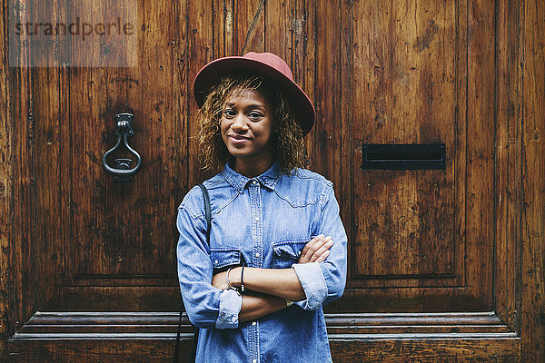 Spanien  Barcelona  Porträt einer lächelnden jungen Frau mit Hut und Jeanshemd vor einer Holztür stehend