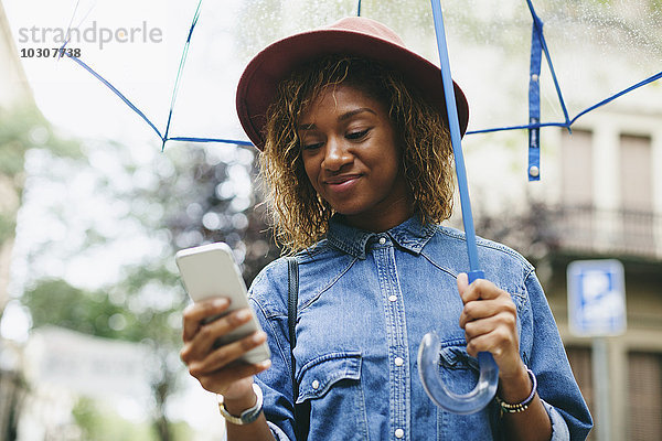 Spanien  Barcelona  Portrait einer lächelnden jungen Frau mit Schirm und Smartphone
