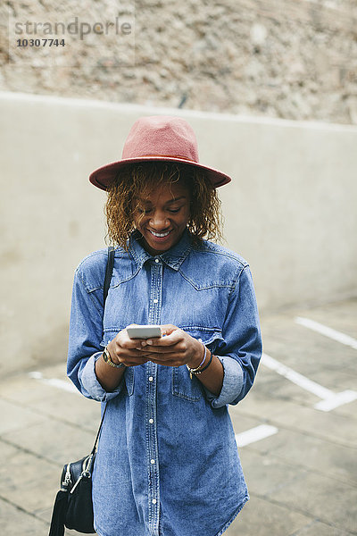 Porträt einer lächelnden jungen Frau mit Hut und Jeanshemd auf dem Smartphone