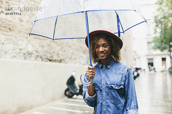 Spanien  Barcelona  Porträt einer lächelnden jungen Frau mit Regenschirm in Hut und Jeanshemd