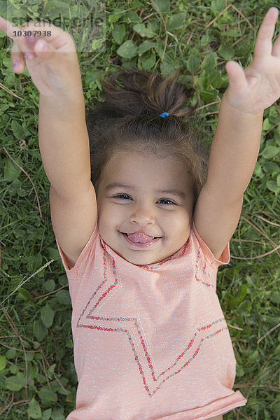 Porträt eines lächelnden kleinen Mädchens  das auf einer Wiese liegt und die Zunge herausstreckt.