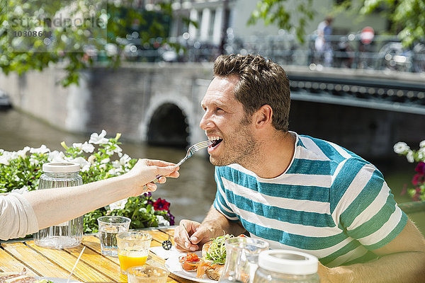 Niederlande  Amsterdam  Frau füttert Mann im Außenrestaurant am Stadtkanal