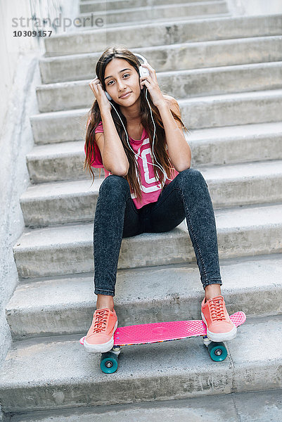 Porträt eines jungen Mädchens  das mit einem Skateboard auf einer Treppe sitzt und Musik mit Kopfhörern hört.