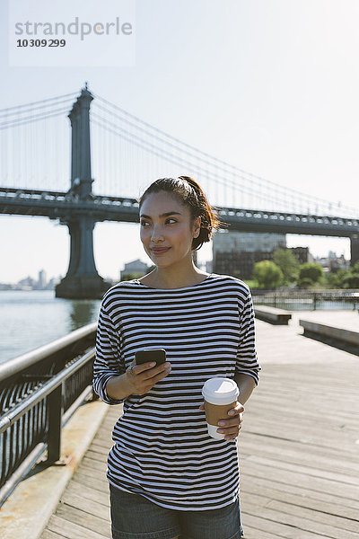 USA  New York City  Porträt einer lächelnden jungen Frau mit Smartphone und Kaffee zum Mitnehmen
