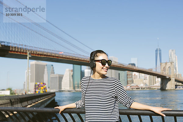 USA  New York City  Portrait einer jungen Frau  die Musik mit Kopfhörern vor der Skyline hört.