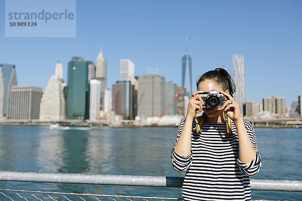 USA  New York City  junge Frau steht vor der Skyline und fotografiert mit der Kamera.