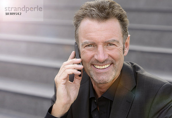 Porträt eines lächelnden Geschäftsmannes  der auf einer Treppe sitzt und mit dem Smartphone telefoniert.
