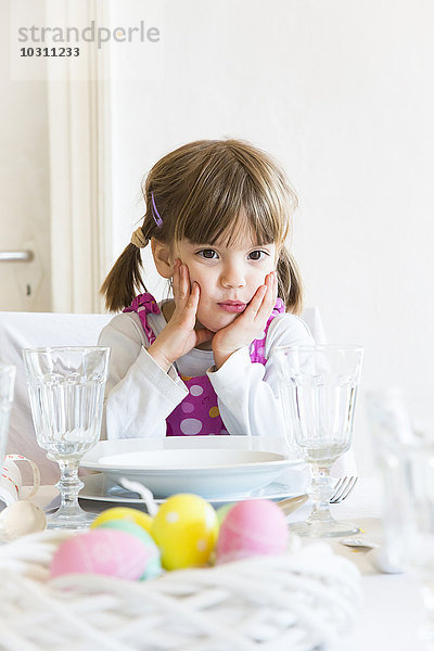 Porträt eines kleinen Mädchens mit Kopf in der Hand  das am gedeckten Esstisch sitzt.