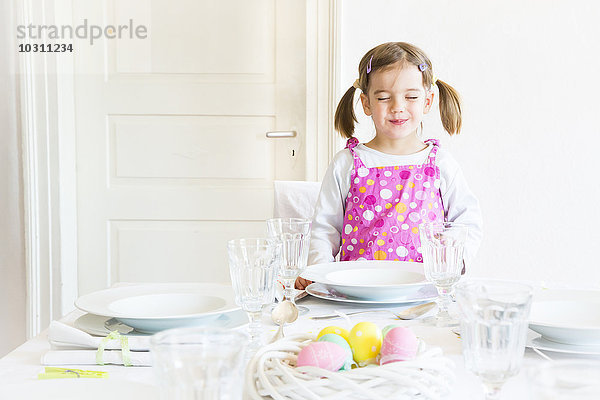 Porträt eines lächelnden kleinen Mädchens mit geschlossenen Augen  das hinter einem gedeckten Tisch steht.