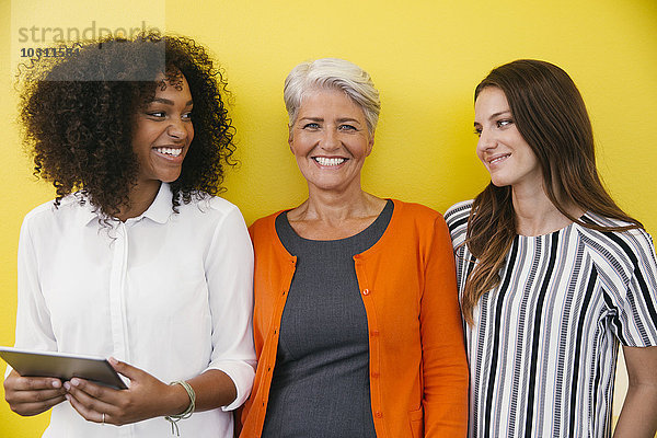 Drei lächelnde Frauen  die vor einer gelben Wand stehen.