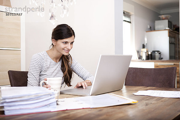 Lächelnde Frau am Holztisch sitzend mit Tasse Kaffee und Laptop