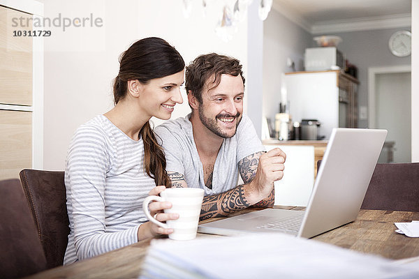 Lächelndes Paar sitzt am Holztisch und schaut auf den Laptop.