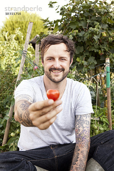 Porträt eines lächelnden Mannes mit Tätowierungen auf den Armen  der im Garten sitzt und Tomaten hält.