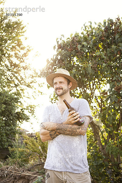 Porträt eines Mannes mit Tätowierungen auf den Armen  der im Garten steht und eine Flasche Bier hält.