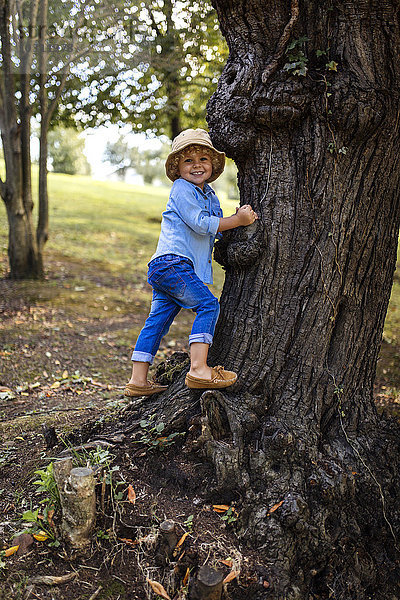 Porträt eines lächelnden blonden Jungen mit Hut neben einem Baum