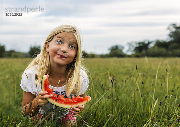 Mädchen mit Wassermelonenscheibe auf einer Wiese stehend und ein Gesicht machend