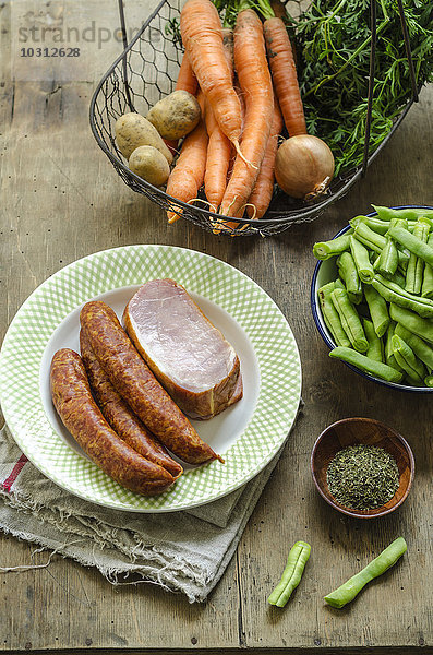 Zutaten für Bohneneintopf  Hackfleischwurst und geräuchertes Schweinekotelett auf Teller  Karotte  Kartoffel  Zwiebel und Kräuter im Korb