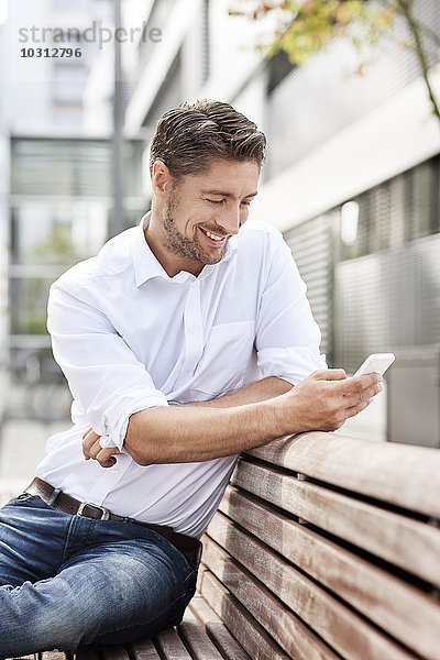 Porträt eines lächelnden Mannes auf einer Holzbank mit Blick auf sein Smartphone