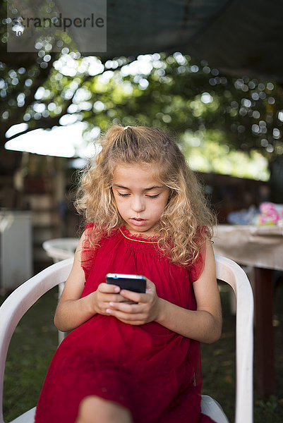 Kleines Mädchen in rotem Kleid auf einem Stuhl sitzend mit Smartphone