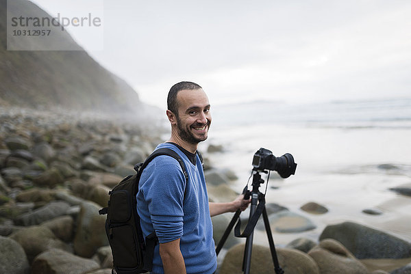 Spanien  Valdovino  Porträt des lächelnden Fotografen am Strand mit Stativ und Kamera