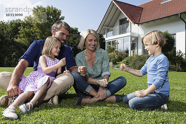Fröhliche Familie sitzt auf Rasen im Garten und spielt Stein-Papier-Schere
