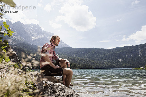 Deutschland  Bayern  Eibsee  lächelnder Mann in Lederhose sitzend am Seeufer